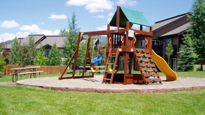 A wooden playground structure with a slide and swings, surrounded by a grassy area and residential buildings in the background. - Direct Unit Access,  Garage, 2 decks, Weber BBQ, Surround Sound Home Theater (Steamboat Springs)
