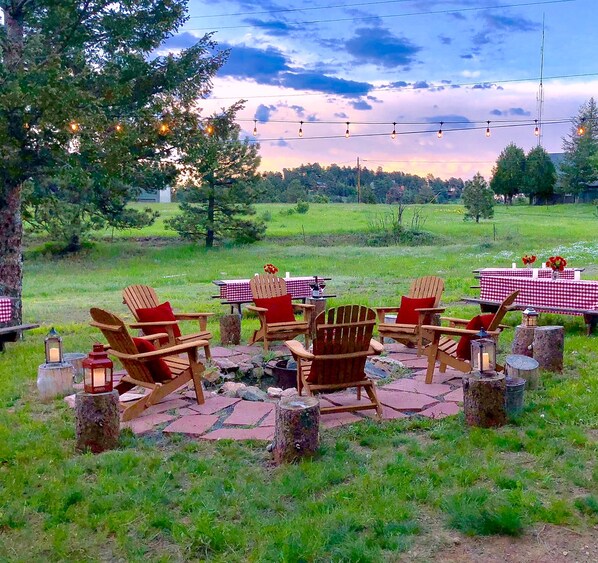An outdoor seating area with Adirondack chairs and checkered tables, surrounded by trees and a grassy field.