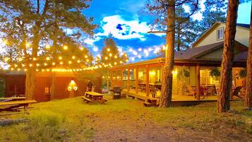 Un espacio comedor rústico al aire libre con luces colgantes, mesas de madera y una zona de estar cubierta.