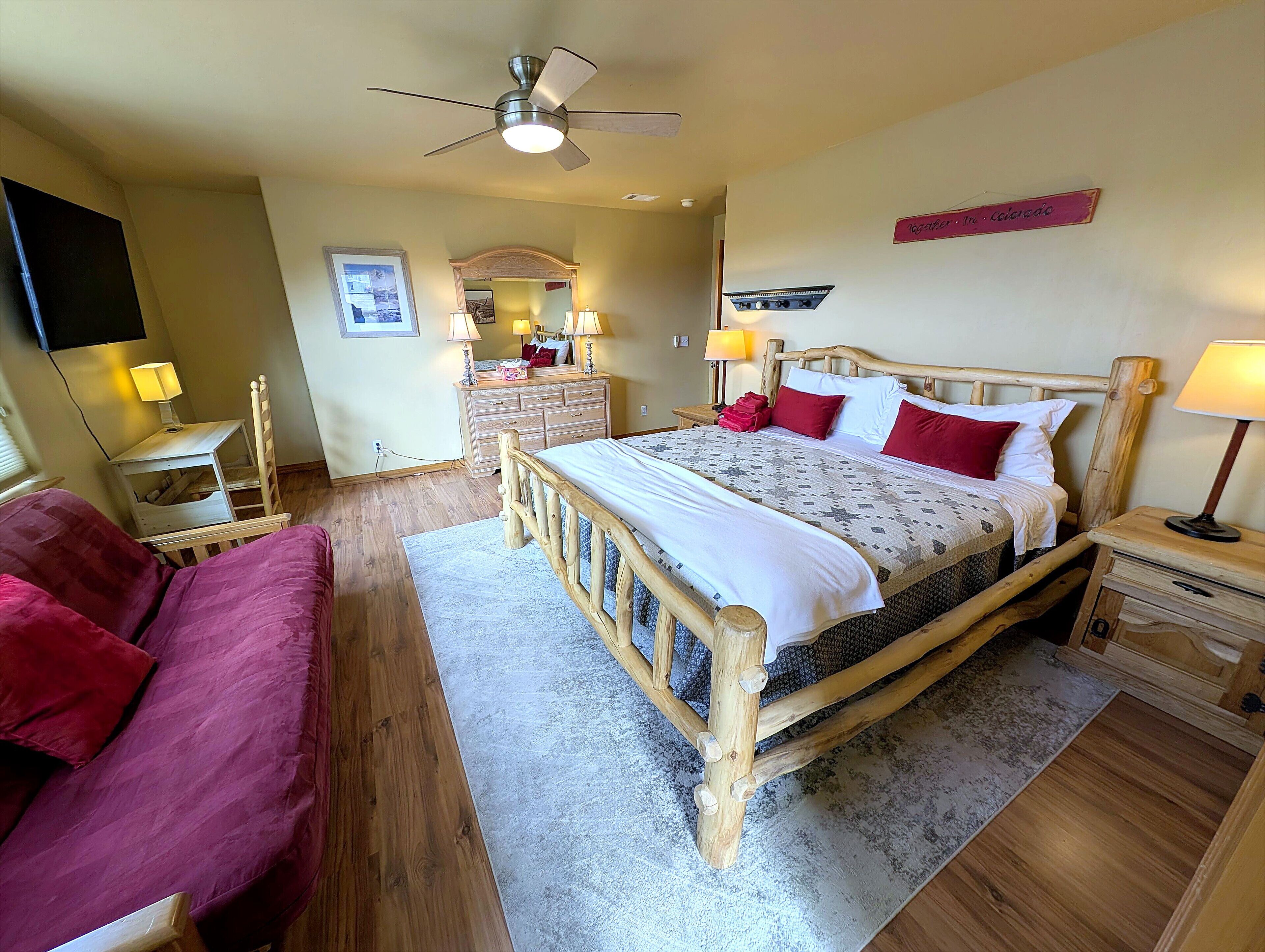 A bedroom with a wooden bed, a desk, a chair, a dresser, a mirror, a ceiling fan, and a flat-screen TV.