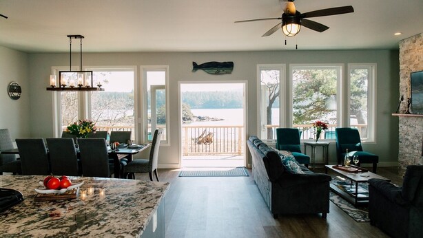 A spacious living area with a large window overlooking a lake, a ceiling fan, and a dining area with a marble countertop. - Custom Waterfront Home near Roche Harbor. Access to Picnic Island. 4-bedrooms. (Friday Harbor)