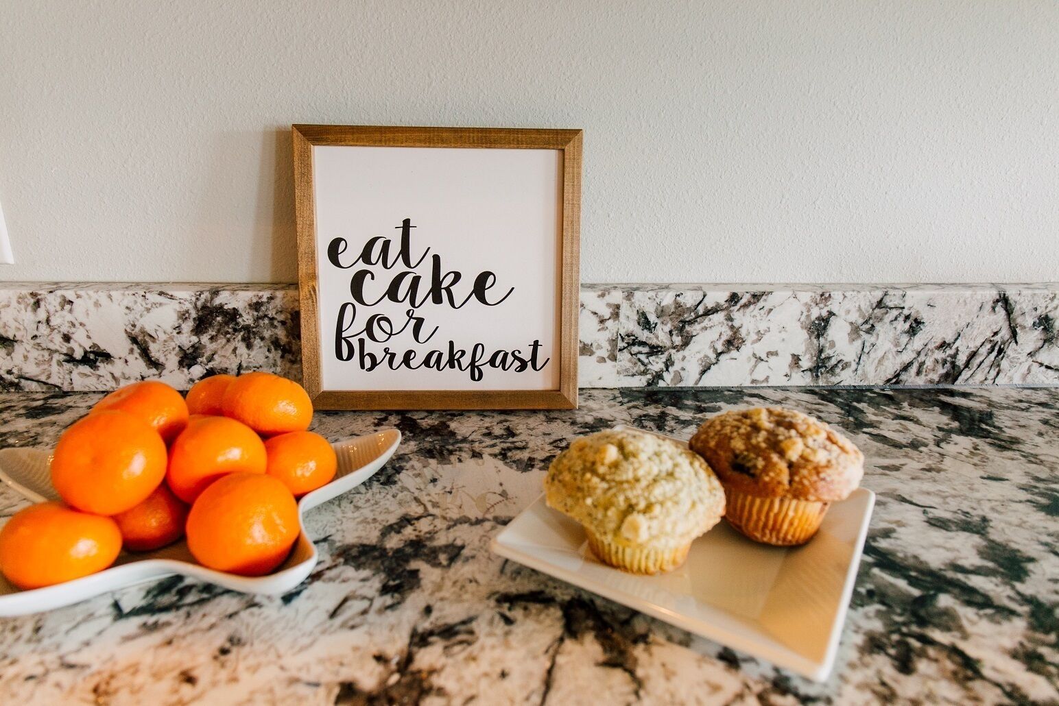 A kitchen counter with a framed sign that reads "eat cake for breakfast," a bowl of oranges, and two muffins.