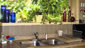 A kitchen with a double sink, a window with a view of greenery, and various kitchen items on the counter. - Paradise Ocean View Private Cottage Solar Power Tropical Flower Gardens (Hana)