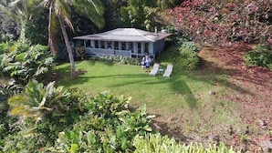 A house with a covered porch, surrounded by lush greenery and a lawn. - Paradise Ocean View Private Cottage Solar Power Tropical Flower Gardens (Hana)