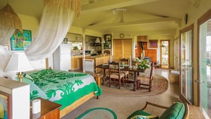 A room with a bed, a dining table, chairs, a kitchen area, and a large window. - Paradise Ocean View Private Cottage Solar Power Tropical Flower Gardens (Hana)