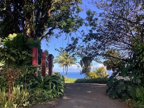 A paved pathway leading to a gate with red pillars, surrounded by lush greenery and palm trees, with a view of the ocean in the background. - Paradise Ocean View Private Cottage Solar Power Tropical Flower Gardens (Hana)