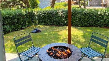 A patio with a circular fire pit table, two metal chairs, and a view of a well-maintained lawn and hedge.