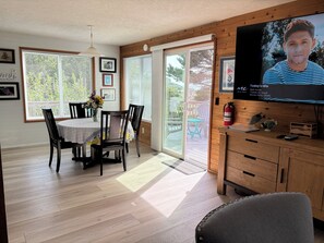 Un área de comedor con una mesa y sillas, un televisor montado en la pared y vistas al exterior a través de amplios ventanales.
