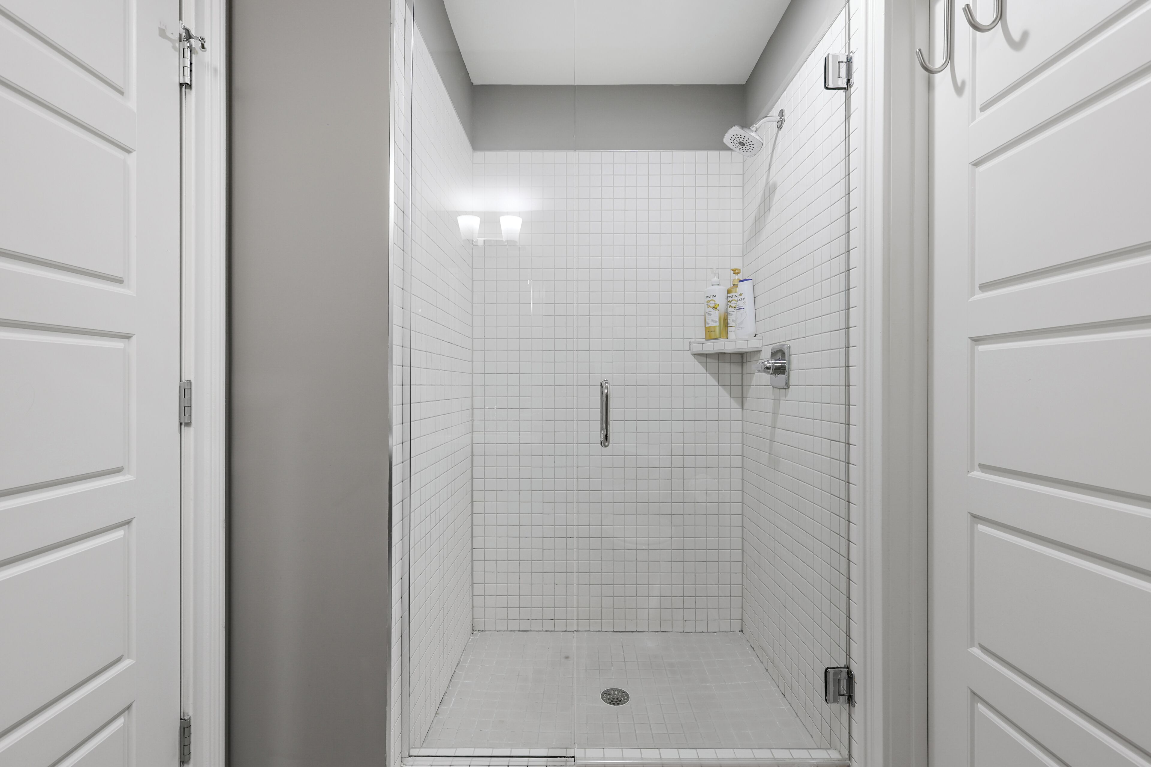 Un box de ducha estrecho con azulejos, una puerta blanca a un lado y un cabezal de ducha sobre el espejo.