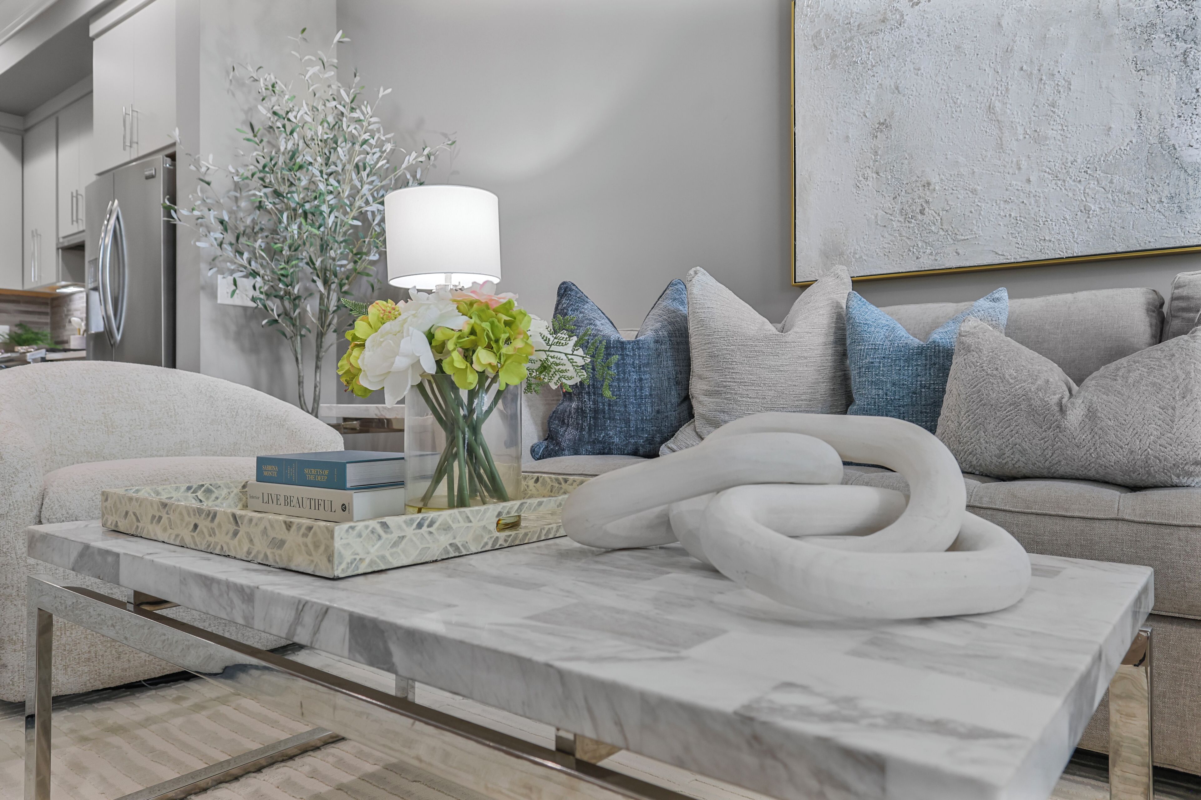 Een moderne woonkamer met een marmeren bijzettafeltje, een bank met decoratieve kussens, een vaas met bloemen en een wandlamp.