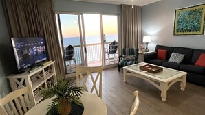 A living room with a television, a dining table with chairs, a sofa, a coffee table, and a balcony with a view of the sea.
