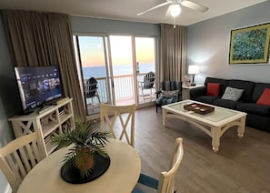 Una sala de estar con televisor, una mesa de comedor con sillas, un sofá, una mesa baja, una planta y un balcón con vista al mar.