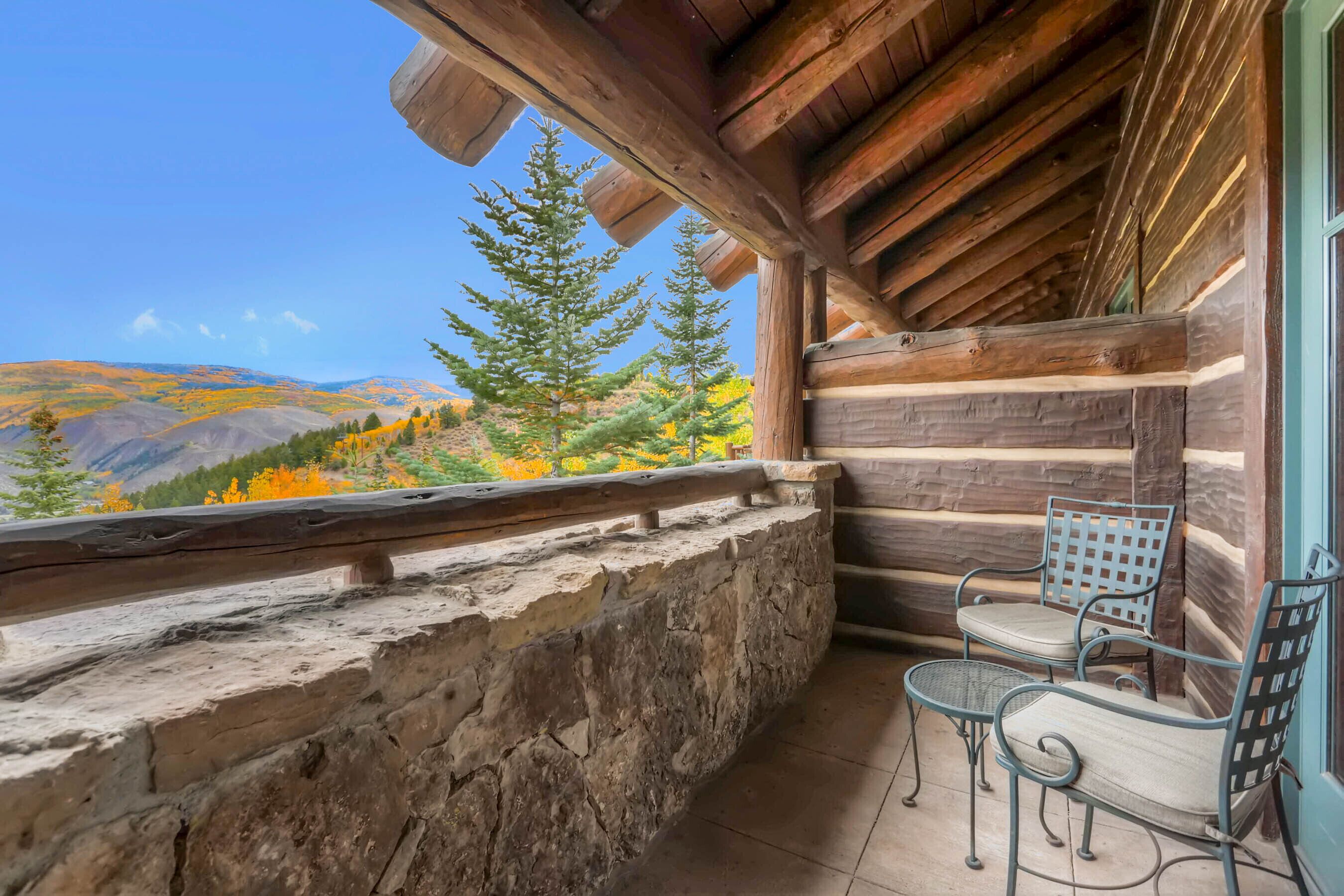 Una cabaña de troncos con pared de piedra y vigas de madera, con dos sillas metálicas y vistas a un paisaje montañoso.