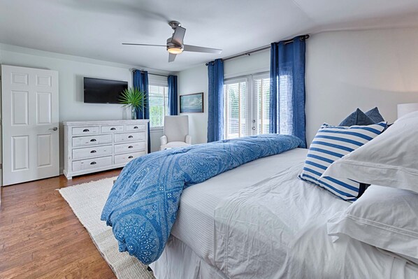 Une chambre à coucher avec un lit, un ventilateur de plafond, une télévision, une commode et une fenêtre avec des rideaux.