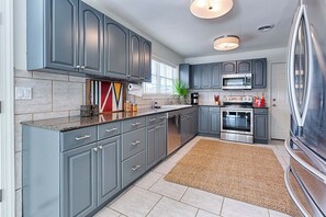 A modern kitchen with grey cabinets, stainless steel appliances, and a woven rug. - Walk to Dining,& the Beach–Chic Cottage, Bikes, Kayak, Golf Cart Available (Santa Rosa Beach)