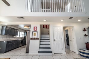 A modern kitchen with dark cabinets and a central island, a staircase leading to a balcony, and a hallway with a door and wall decorations. - Walk to Dining,& the Beach–Chic Cottage, Bikes, Kayak, Golf Cart Available (Santa Rosa Beach)