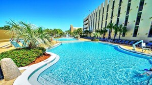 A swimming pool with lounge chairs and palm trees, surrounded by a building with multiple floors and balconies.