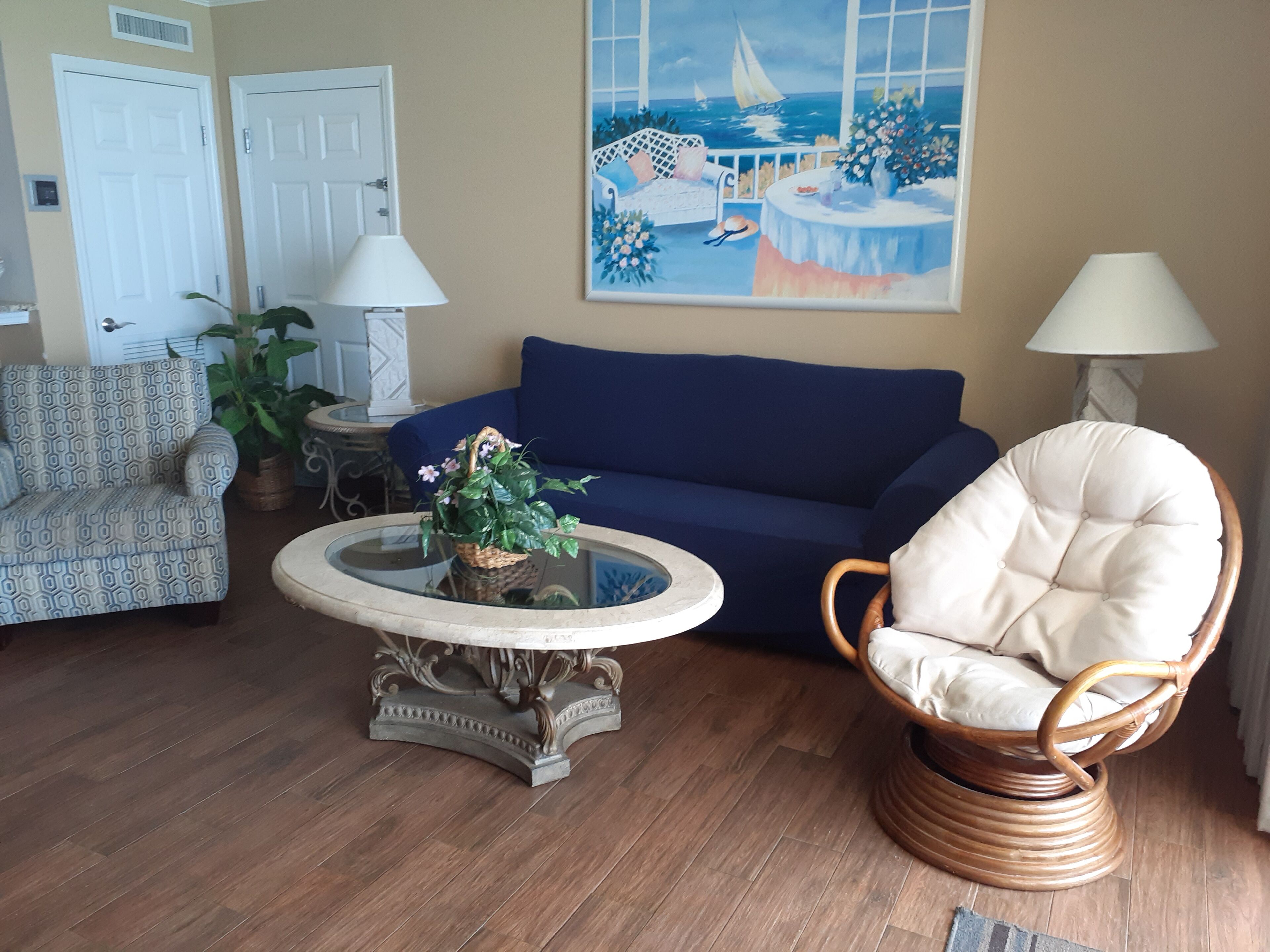 A living room with a blue sofa, a wicker chair, a glass coffee table with a plant, and a painting of a seaside scene.