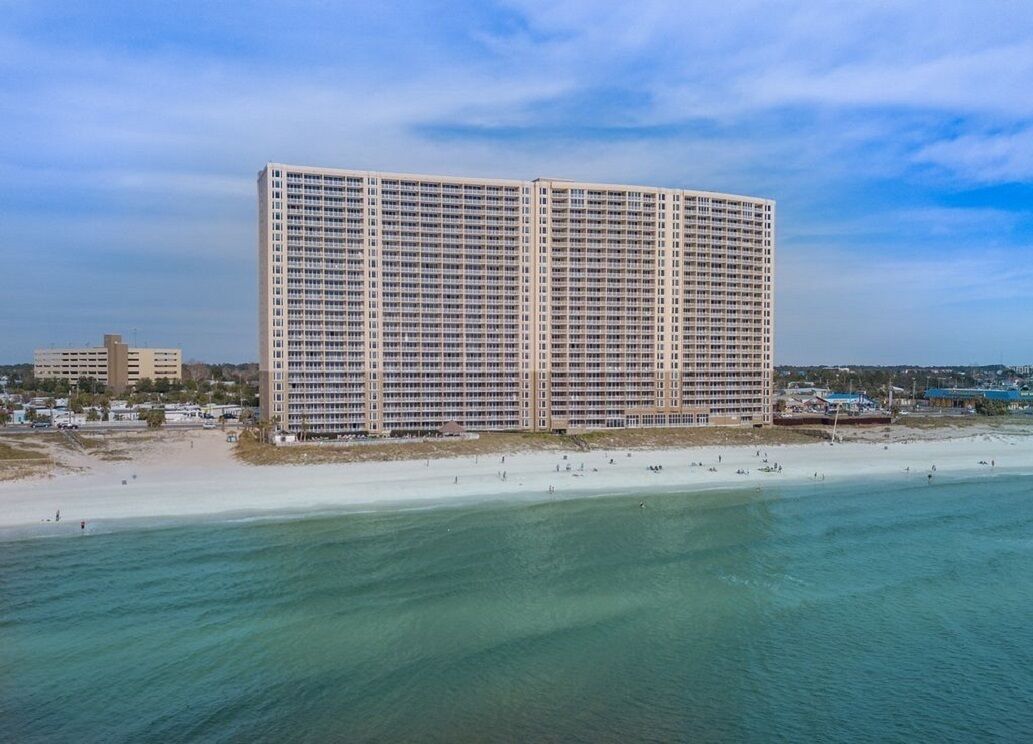 A large, rectangular hotel with multiple floors, situated near a beach with a sandy shore and clear blue water.