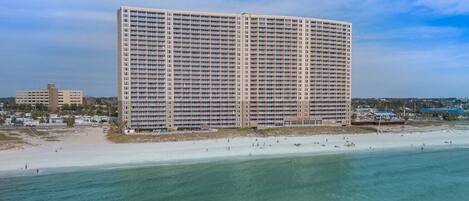 A large, rectangular hotel with multiple floors, situated near a beach with a sandy shore and clear blue water.