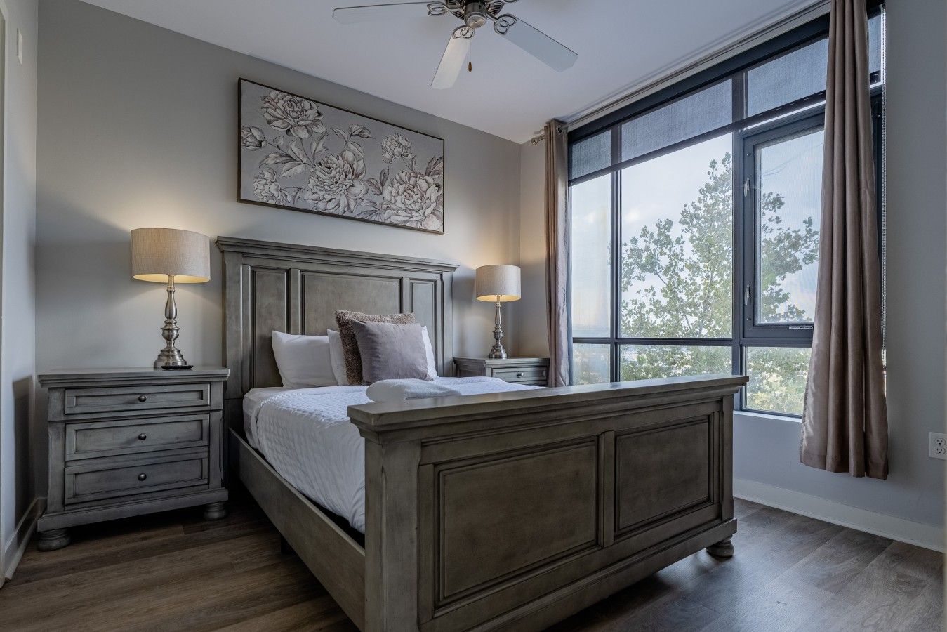 A bedroom with a large bed, two bedside tables with lamps, a ceiling fan, and a window with curtains.