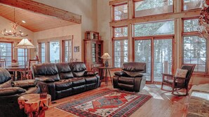 A spacious living room with a leather sofa, two armchairs, a rocking chair, a patterned rug, and large windows offering a view of the forest. - Rivamonte by Cottonwood Lodging (Brighton)