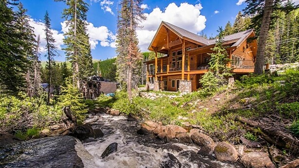 A log cabin with a stone foundation, surrounded by dense forest and a flowing stream. - Rivamonte by Cottonwood Lodging (Brighton)