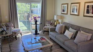 A living room with a grey sofa, a glass coffee table, a small side table with a vase of flowers, and a view of the outdoors through large windows. - Carmel-by-the-Sea Escape (Carmel-By-The-Sea)