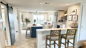 A modern kitchen with a central island, bar stools, and a dining area with a view. - Tropical Island Getaway Beachfront Gulf Views-Pool-Ferry & Grocery Delivery incl (Placida)