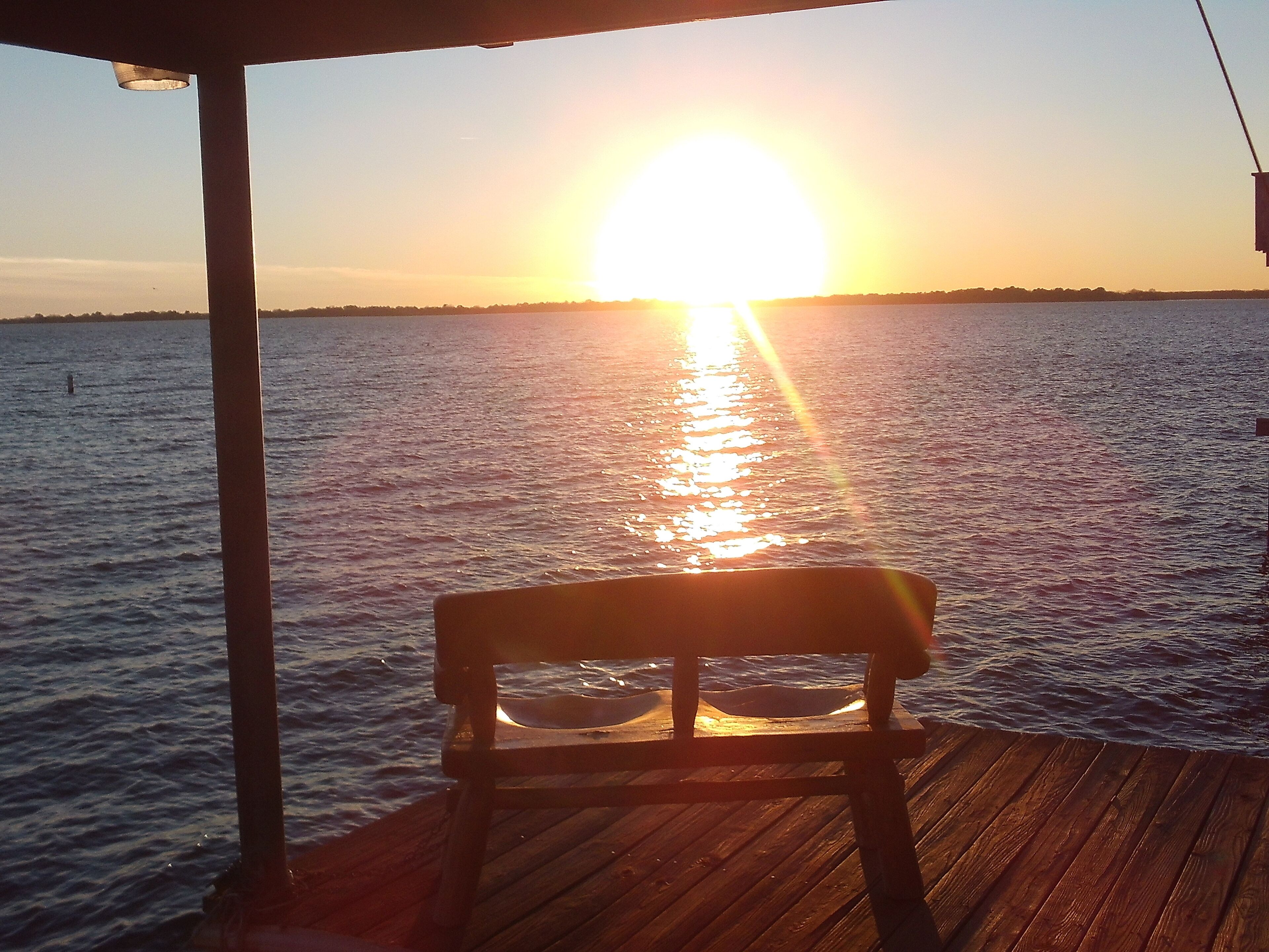 A wooden deck with a bench facing a sunset over water.