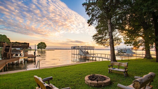 A lakeside deck with wooden chairs, a fire pit, and a dock extending into the water.