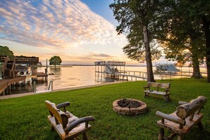 A lakeside deck with wooden chairs, a fire pit, and a dock extending into the water.