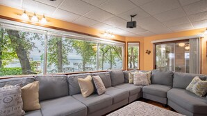 A modern living room with a grey sectional sofa, patterned ottoman, and large windows offering a view of the outdoors.