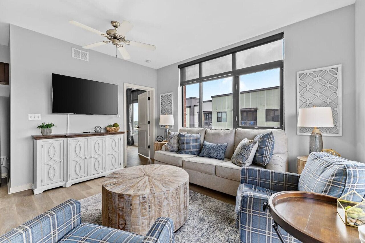 A modern living room with a large window, a ceiling fan, a flat-screen TV, and a sofa with plaid cushions.