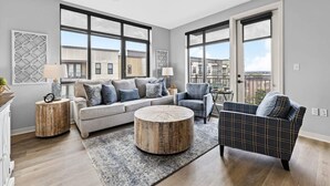 Un salon spacieux avec un canapé gris, un fauteuil à motifs et une table basse ronde. De grandes fenêtres offrent une vue sur la ville.