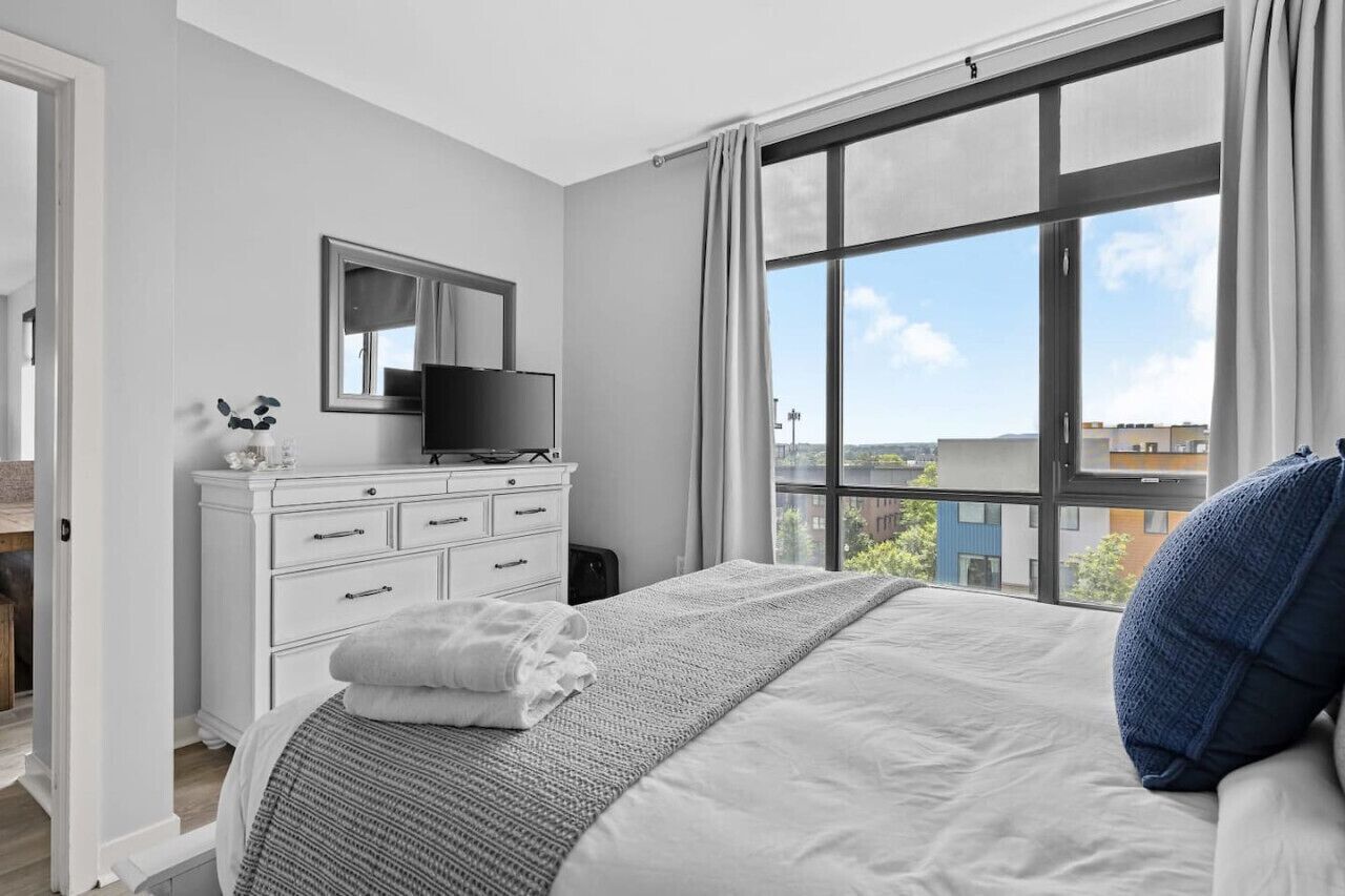 Ein modernes Schlafzimmer mit großem Fenster, einem Flachbildfernseher, einer weißen Kommode und einem ordentlich bezogenen Bett.