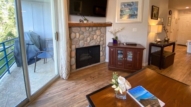 A living room with a stone fireplace, a flat-screen TV mounted above it, a wooden coffee table with a vase of flowers, and a sliding glass door leading to a balcony. - Inn Building Condo #206 Beautiful updated condo (Garden City)