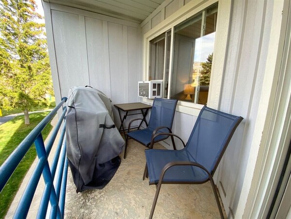 Un balcon abrité avec une table et deux chaises, un barbecue couvert et une fenêtre avec des stores.