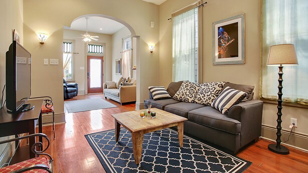 A living room with a sofa, coffee table, and a television. There is a door leading to another room. - 2-3 N stays - Spacious and Comfy, Walk to Whole Foods, bars and shops, sleeps 12 (New Orleans)