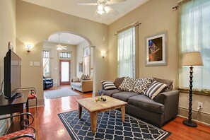 A living room with a sofa, coffee table, and a television. There is a door leading to another room. - 2-3 N stays - Spacious and Comfy, Walk to Whole Foods, bars and shops, sleeps 12 (New Orleans)