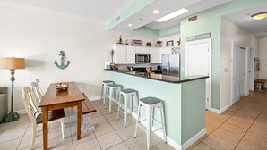 A kitchen with a dining area, white cabinets, a stainless steel refrigerator, and a sign that reads "COAST ALONG". - Indies 704 | 3BR/2BA Penthouse Condo | Sleeps 9 | Gulf Views & Heated Pool (GULF SHORES)