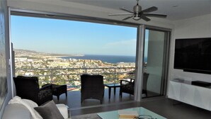 Una sala moderna con sofá, sillas, una mesa de centro y televisor. Cuenta con un gran ventanal corredizo que da a un balcón con vistas a la ciudad.