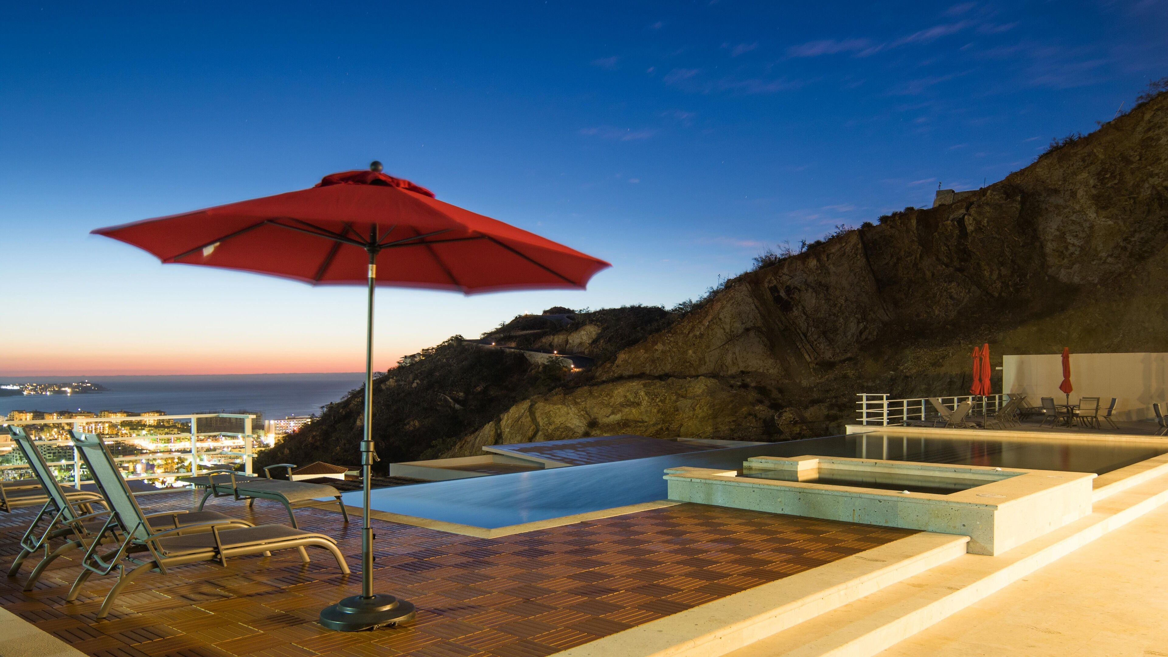 Une piscine à débordement située sur le toit, avec un parasol rouge, des chaises longues et une vue sur la ville au crépuscule.