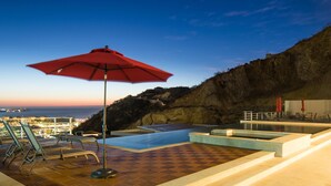 Piscina infinita en la azotea con una sombrilla roja, sillas de descanso y vistas de la ciudad al anochecer.