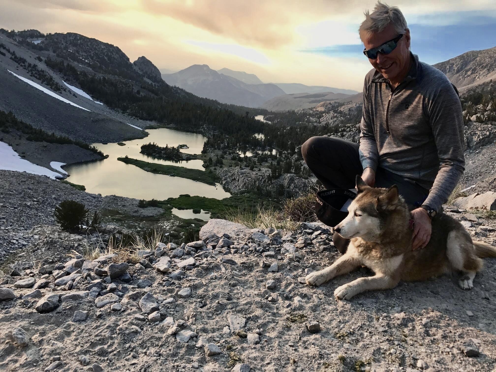 Una persona sentada sobre terreno rocoso con un perro, contemplando un paisaje montañoso con un lago.