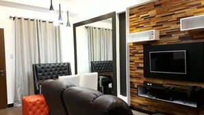 A modern living room with a leather sofa, a flat-screen TV mounted on a wooden wall, and a large mirror. - N603 Outlook Ridge Residences (Baguio)
