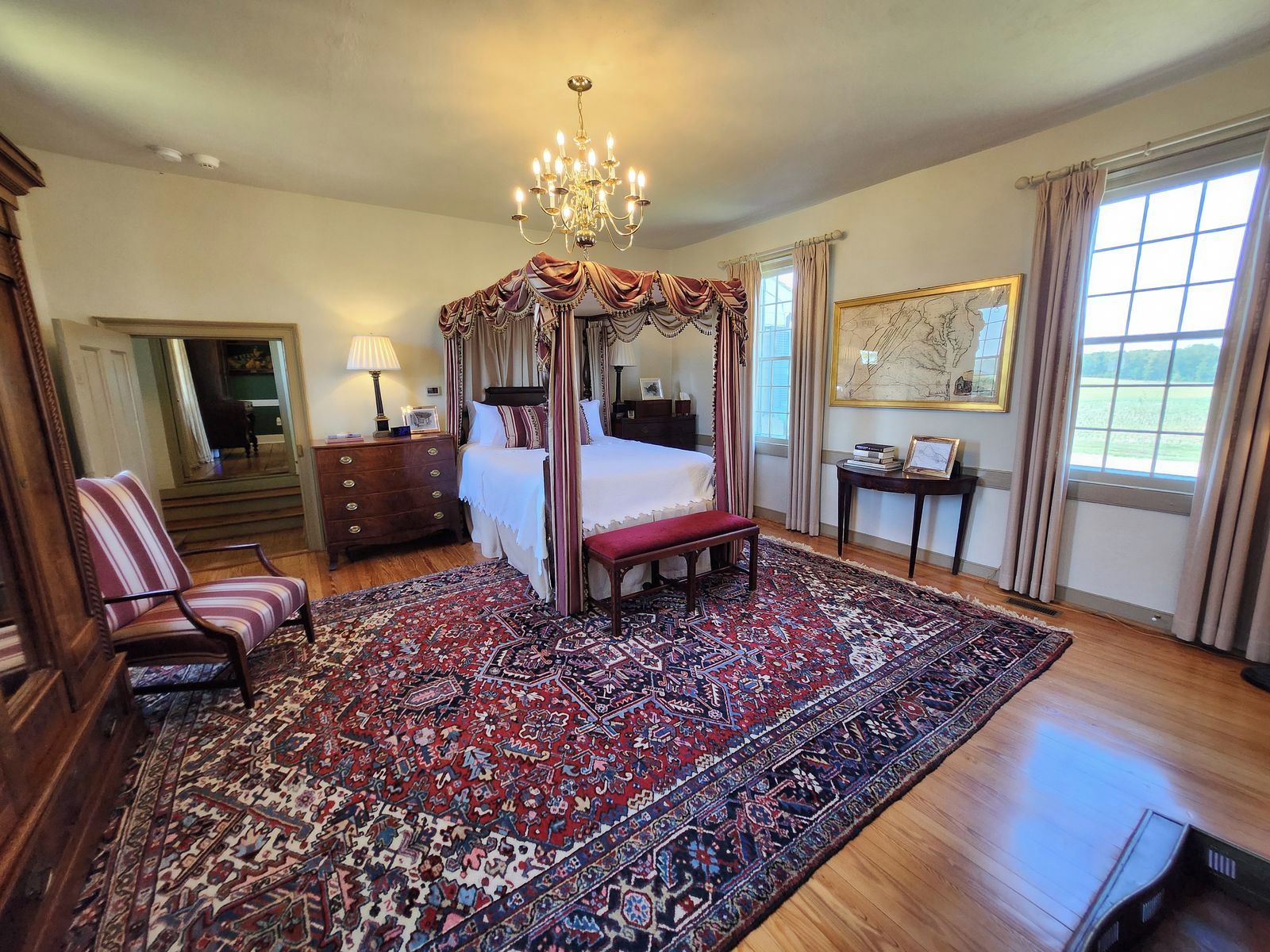 A four-poster canopy bed with a red and white patterned rug, a wooden bench, a chair, and a large window with curtains.