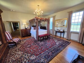 A four-poster canopy bed with a red and white patterned rug, a wooden bench, a chair, and a large window with curtains.