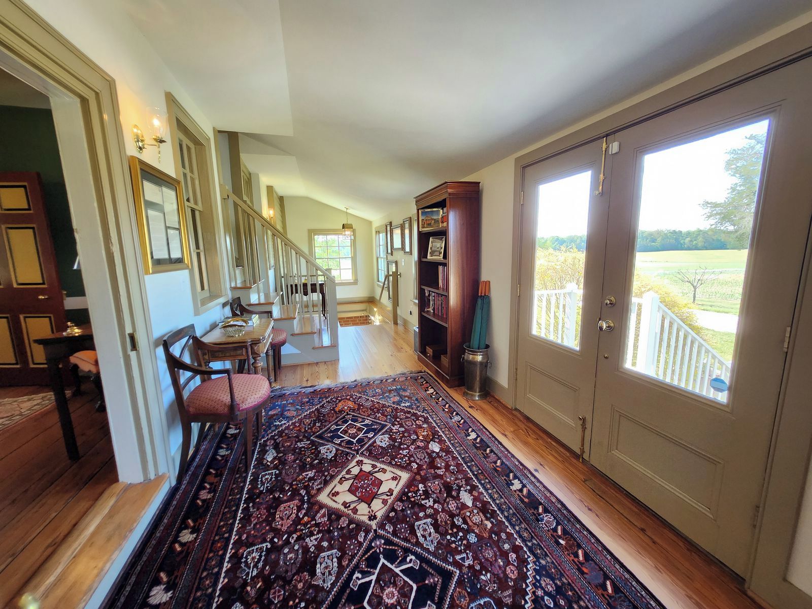 A spacious hallway with a wooden floor, a dining area with a table and chairs, and a large window offering a view of a green landscape.