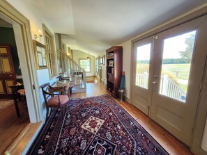 A spacious hallway with a wooden floor, a dining area with a table and chairs, and a large window offering a view of a green landscape.